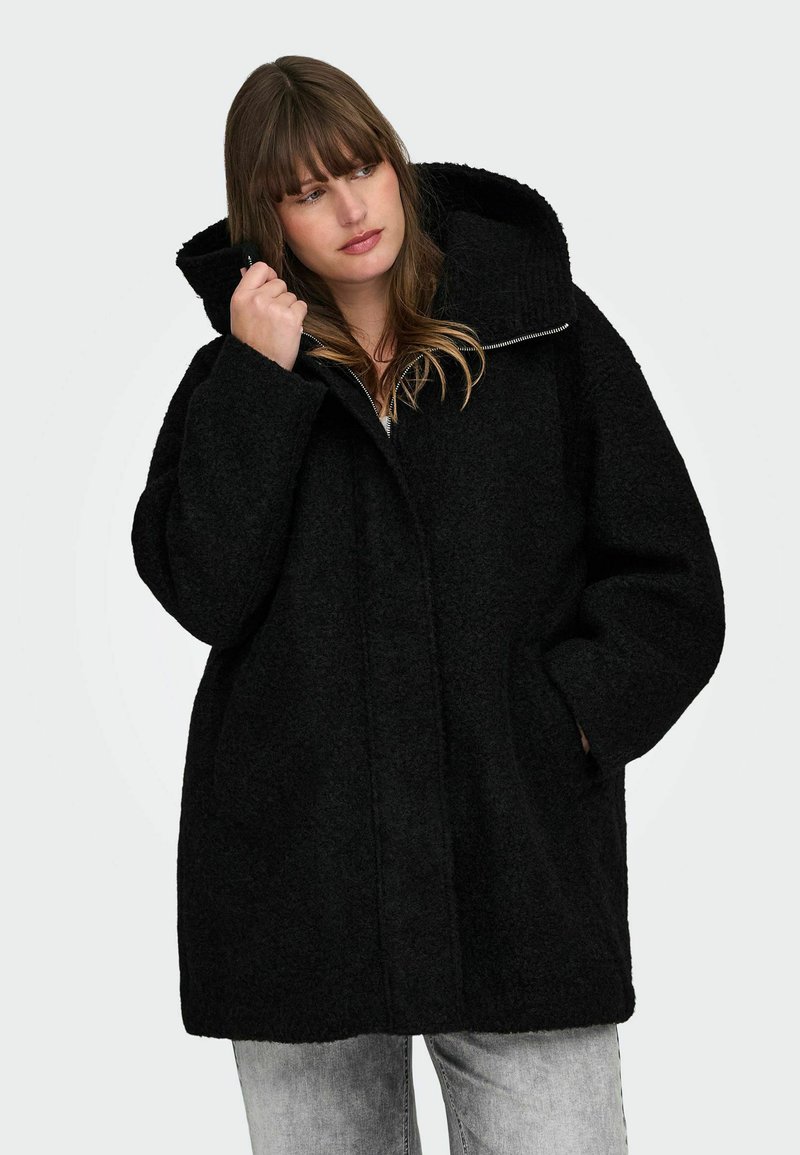 Črn, teksturiran, prevelik plašč z visokim ovratnikom, zadrgo in velikim kapuco. Ima stranske žepe in ohlapen, sproščen kroj.