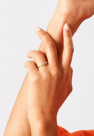Anello d'oro con una piccola pietra preziosa trasparente, indossato su una mano con un tono di pelle naturale, dita leggermente incrociate. Banda metallica liscia, design minimale.
