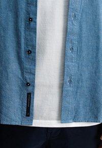 Lichtblauw chambray overhemd met zwarte knopen en label; gelaagd over een wit T-shirt, gecombineerd met marineblauwe broek. Zachte textuur, casual ontwerp.