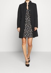 Black wool coat with a high collar and button closure, worn over a black dress with a white and peach floral pattern.