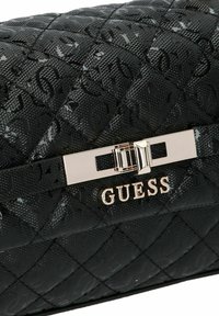 Sac à main noir matelassé avec motif texturé, fermeture à verrou en métal argenté et logo "GUESS" argenté sur le rabat avant.