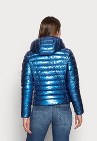 Vrouw met een glanzende blauwe doorgestikte jas met capuchon en blauwe spijkerbroek, van achteren weergegeven tegen een effen lichte achtergrond.