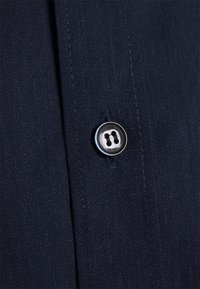 Un singolo bottone bianco e nero cucito su una stoffa in tweed blu scuro, con cuciture visibili.