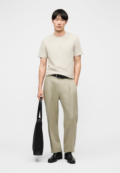 Άνδρας που φοράει μπεζ μπλουζάκι και παντελόνι, μαύρη ζώνη και παπούτσια, κρατώντας μια μαύρη τσάντα ώμου με το ένα χέρι στην τσέπη, πάνω σε απλό φόντο.