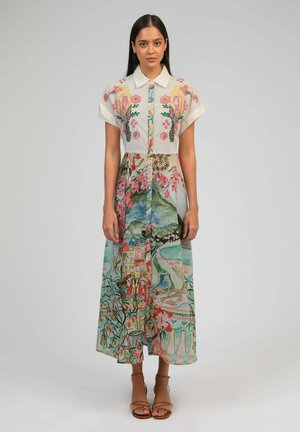 Femme debout, portant une robe à col boutonnée à manches courtes avec des motifs floraux colorés et des paysages, ainsi que des sandales à bride à la cheville.