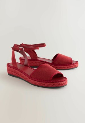 Raudoni espadrilės tipo sandalai su atvira pirštine, minkšta zomšos viršūne, pintomis džutų tarpuliu ir reguliuojama blauzdos juosta su metaline sagtimi.