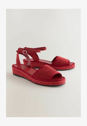 Raudoni espadrilės tipo sandalai su atvira pirštine, minkšta zomšos viršūne, pintomis džutų tarpuliu ir reguliuojama blauzdos juosta su metaline sagtimi.