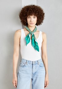 Green patterned silk scarf tied around the neck, featuring geometric designs and smooth texture. Worn with a white tank top and high-waisted jeans.