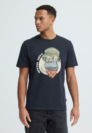 Jonge man in een zwart t-shirt met een afbeelding van een nijlpaard met een emmerhoed, zonnebril en bandana, met het label "Hipster Hippo", staande tegen een witte achtergrond.
