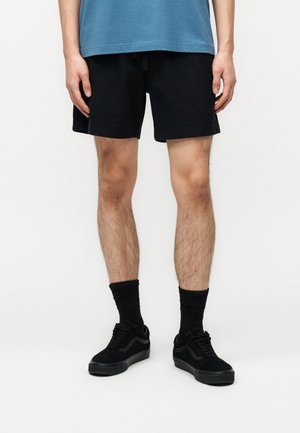 Mann trägt schwarze Shorts, schwarze Socken und schwarze Turnschuhe, steht vor einem schlichten hellen Hintergrund.