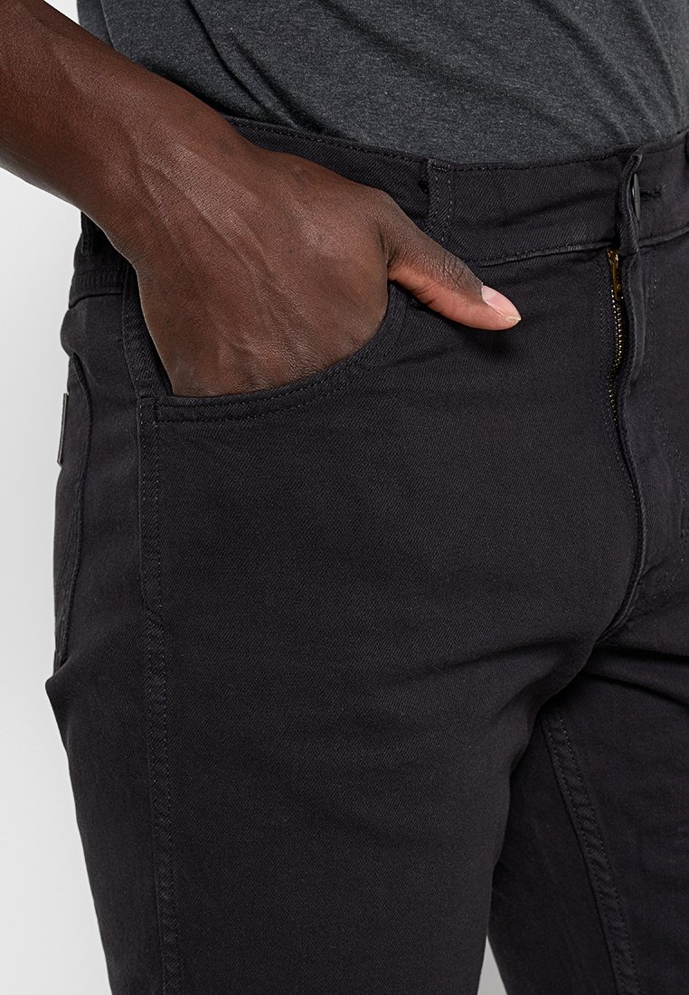 Czarne dżinsy mają klasyczny prosty krój, staranne przeszycia, przednie kieszenie i zamek błyskawiczny. Tkanina wydaje się trwała i teksturowana.