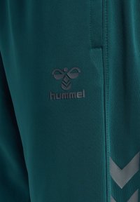 Türkise Sporthose aus glattem, leichtem Material. Mit einem auffälligen schwarzen "hummel"-Logo und grauen Chevron-Akzenten an der Seite.