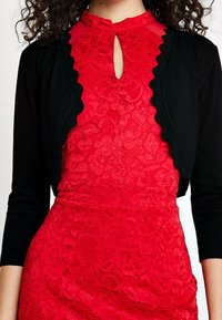 Rotes Spitzenkleid mit einem gewellten Ausschnitt und transparenten Akzenten, kombiniert mit einer enganliegenden schwarzen Strickjacke. Beide Teile haben angepasste Ärmel.