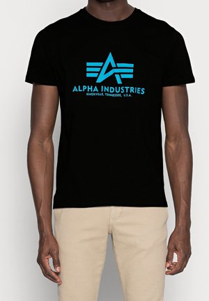 Osoba ubrana w czarną koszulkę z niebieskim logo "Alpha Industries Knoxville, Tennessee, U.S.A." i beżowe spodnie, stojąca na jednolitym tle.