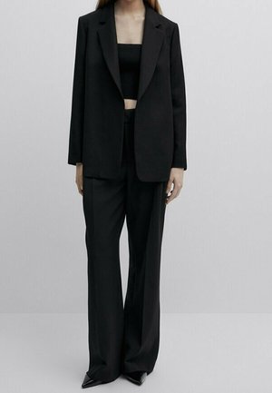 Kvinde iført en sort oversized blazer, sorte bukser med brede ben, sort crop top og sorte spidse sko mod en ensfarvet baggrund.