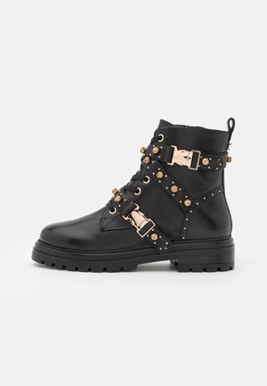 Schwarzer Leder-Stiefelette mit goldenen dekorativen Schnallen, Nieten und einer klobigen Sohle.