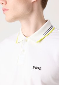 Fehér meglapú ing, sárga díszítéssel a gallérnál, fekete "BOSS" felirattal és szürke csíkokkal a gallér széle mentén.