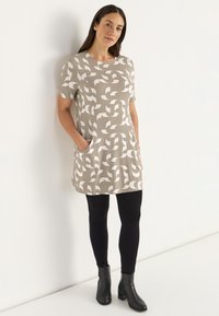 Beige kurzärmliges Kleid mit einem weißen eingewebten Muster, ausgestattet mit zwei Taschen. Kombiniert mit schwarzen Leggings und Stiefeletten.