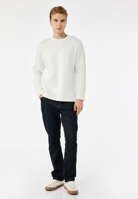 Junger Mann mit Ohrringen, der ein weißes langärmliges Hemd, dunkle Jeans und weiße Sneaker trägt. Er steht mit einer Hand in der Tasche vor einem weißen Hintergrund.
