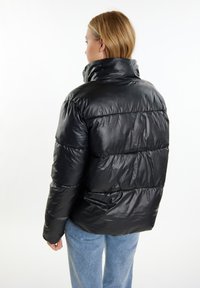 Jachetă puffer neagră cu guler înalt, finisaj lucios și detalii cu cusături orizontale. Purtați cu pantaloni ușor albaștri.