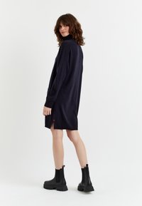 Abito a maglia blu navy con colletto alto, spacchi laterali e cuciture di contrasto rosse. Abbinato a stivaletti neri alla caviglia con suola spessa.