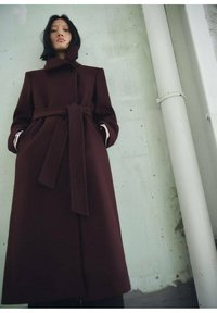 Manteau en laine bordeaux avec une taille ceinturée, un col haut et deux poches latérales, présentant des revers intérieurs contrastants. Texture lisse, design longueur genou.