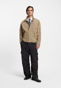 Ταμπά σακάκι από σουέτ με όρθιο γιακά, κλείσιμο με φερμουάρ και πλαϊνές τσέπες· συνδυασμένο με μαύρο παντελόνι cargo και μαύρα παπούτσια.