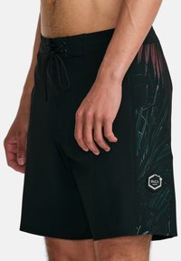 Black board shorts featuring a green and maroon tropical leaf pattern on one side; includes a drawstring waistband and logo patch detail.