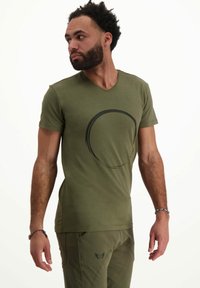 Vihreä oliivi puuvillapaita, jossa on V-pääntie ja etuosassa musta pyöreä graafinen kuvio. Pehmeä tekstuuri, istuva leikkaus ja lyhyet hihat.