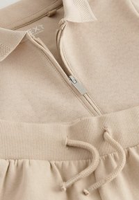 Top zippé en tricot beige avec col, associé à un pantalon à taille élastique côtelé avec cordon de serrage, montrant de près la texture et les détails.