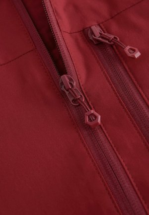 Nahaufnahme einer roten Stoffjacke mit einem doppelten Reißverschluss, einer strukturierten Oberfläche und einem sechseckigen Reißverschlussanhänger. Nahtdetails entlang der Reißverschlusslinie sichtbar.