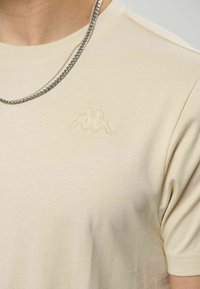 Beige bomulls-t-skjorte med en liten, ton-i-ton brodert logo på brystet, supplert med et sølvkjede.