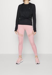 Musta pitkähihainen paita, jossa on solmittu yksityiskohta vyötäröllä, yhdistettynä vaaleanpunaisiin leggings-housuihin, joissa on verkkoaukkoja. Keveät urheilukengät.