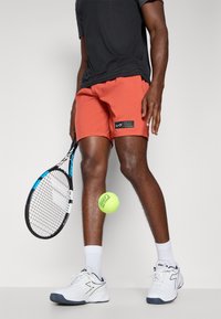Rot-orange Tennis-Shorts mit einem schwarzen Logo-Patch, kombiniert mit weißen Sportschuhen und einem schwarzen T-Shirt. Tennisschläger und Ball sind enthalten.