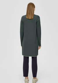 Robe pull longue à manches longues de couleur vert foncé avec des fentes sur les côtés, un col rond et des poignets côtelés. Assortie avec un pantalon sombre et des baskets.