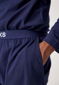 Marineblauwe katoenen loungewear met een brede elastische tailleband met de tekst "KKS", zijzakken en een gladde textuur. Close-up van de taille en hand.