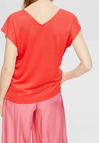 Koraliowy top w serek z krótkimi rękawami, wykonany z gładkiego materiału. Połączony z jasnoróżowymi spodniami typu wide-leg, o luźnym kroju.