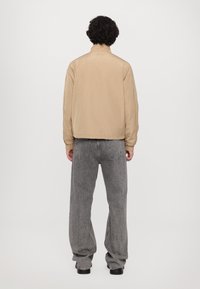 Beige lett jakke med oppreist krage, elastiske mansjetter og rett snitt, kombinert med løse grå jeans og svarte sko.