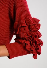 Punainen neulepaita, jossa teksturoidusta neuloksesta valmistettu kangas, pitkät hihat, joissa kerrokselliset röyhelökoristeet, ja resorit hihansuissa. Lähikuva vasemman käden yksityiskohdasta.