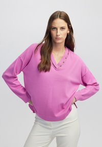 Roze brei-sweater met een diepe V-hals en subtiele decoratieve accenten, voorzien van lange mouwen en een relaxed fit. Gecombineerd met witte broek.