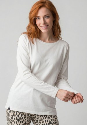 Lächelnde rothaarige Frau richtet den Ärmel eines weißen Langarmshirts, trägt Leopardenmuster-Hose und steht vor einem schlichten grauen Hintergrund.