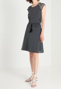 Robe noire à motifs avec des manches courtes et une taille cintrée. Le tissu est léger et présente un design géométrique. Associée à des sandales blanches.