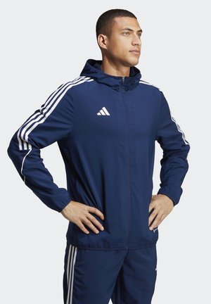 Marine sportjack met witte accenten en drie verticale strepen op de mouwen. Voorzien van een ritssluiting en een verstelbare capuchon. Soepele textuur.