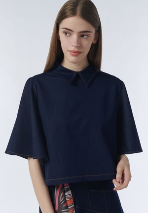 Jonge vrouw met lang bruin haar die een marineblauwe oversized top draagt met wijde mouwen en een kraag, kijkt iets naar links.