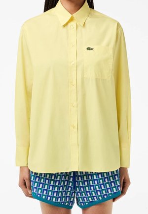 Personne portant une chemise boutonnée à manches longues jaune clair avec une poche poitrine et un logo crocodile vert, associée à un short bleu à motif géométrique.