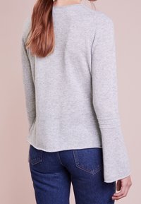 Frau mit langen roten Haaren trägt ein hellgraues Langarmoberteil und dunkle blaue Jeans, von hinten gegen einen neutralen Hintergrund gezeigt.