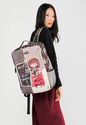Junge Frau trägt ein schwarzes Oberteil und maroonfarbene Hosen, während sie einen bunten Rucksack mit Cartoon- und Paris-Motiven trägt.