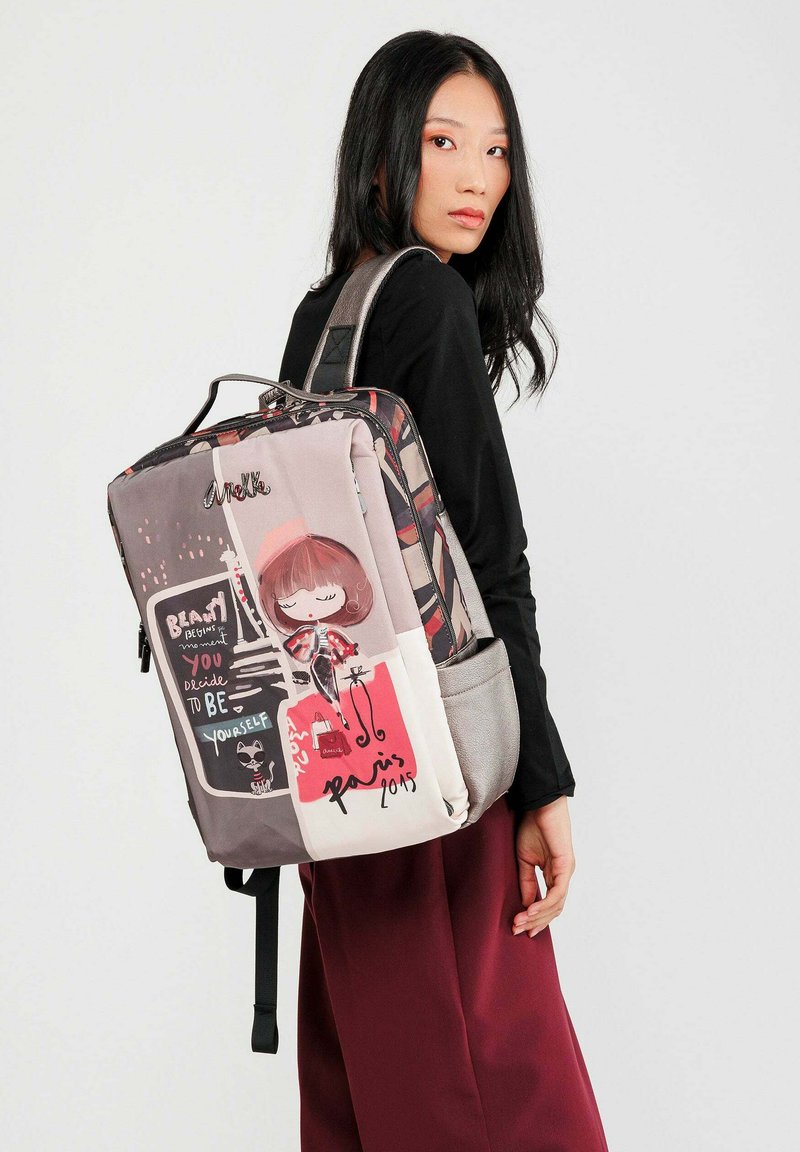 Junge Frau trägt ein schwarzes Oberteil und maroonfarbene Hosen, während sie einen bunten Rucksack mit Cartoon- und Paris-Motiven trägt.