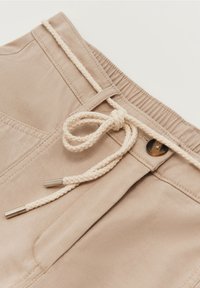 Shorts en tissu beige avec cordon de serrage en corde tressée, présentant une extrémité en métal et une fermeture à bouton à la taille. Texture lisse et coutures.