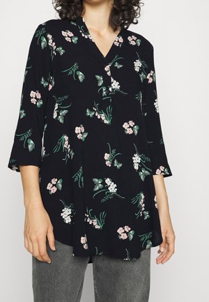 Donna che indossa un blouse nero a maniche lunghe con stampa floreale e di farfalle in rosa e bianco, abbinato a jeans grigi.